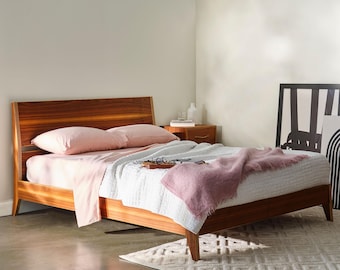 May include: A wooden bed frame with a headboard, featuring a light pink sheet set, white quilted comforter, and a pink throw blanket. The bed is in a well-lit room with a small wooden nightstand and a decorative rug.