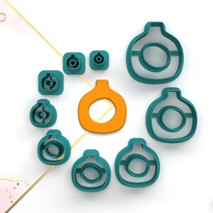 Puede incluir: Un conjunto de cortadores de galletas de color azul turquesa en varias formas, incluyendo una forma redonda con un círculo más pequeño en el interior, una forma de lágrima y una forma cuadrada. Los cortadores están dispuestos sobre una superficie blanca. También hay un solo cortador de galletas naranja en forma de calabaza.