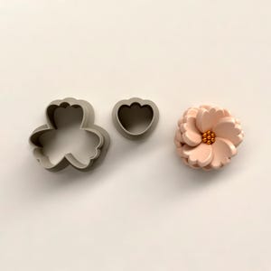 Puede incluir: Tres elementos con temática floral: dos cortadores de galletas grises y una flor color melocotón. Los cortadores de galletas tienen forma de flor y de corazón. La flor tiene múltiples pétalos y un centro dorado.