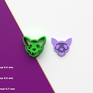 Op de afbeelding: Twee 3D-geprinte koekjesvormen in de vorm van een katten schedel, een groene en een paarse. De vormen worden op een witte achtergrond getoond. De tekst "Sharp 0.4 mm", "Sharp 0.6 mm" en "Normal 0.7 mm" is zichtbaar onder de vormen.