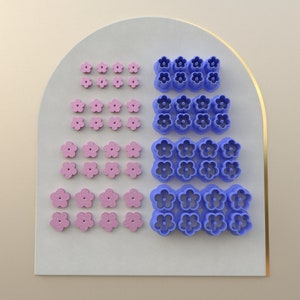 Puede incluir: Un juego de 40 cortadores de galletas con forma de flor, 20 de color rosa y 20 de color azul. Los cortadores están dispuestos en filas sobre una superficie blanca.