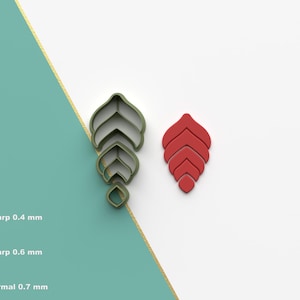 Puede incluir: Dos cortadores de galletas impresos en 3D, uno verde y otro rojo, con forma de hoja estilizada de múltiples capas. Los cortadores se muestran sobre una superficie blanca con un fondo verde y dorado.