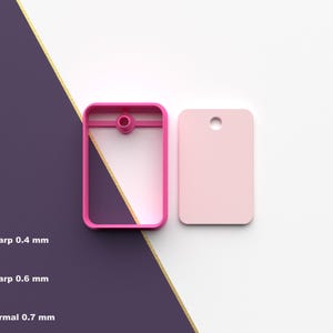 Puede incluir: Un objeto rectangular rosa con un agujero y un marco rosa a juego. El marco tiene una pequeña protuberancia circular. El objeto está al lado de una etiqueta rectangular rosa con un agujero. El fondo es una división de morado y blanco con una franja dorada.