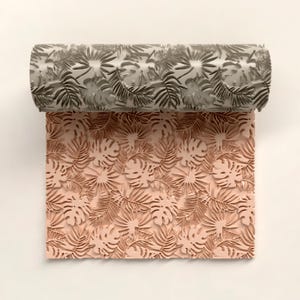 Puede incluir: Un rodillo de pintura gris con un diseño de hojas tropicales repetidas, enrollado sobre una sección de papel rosa. El papel muestra el mismo patrón de hojas en relieve.