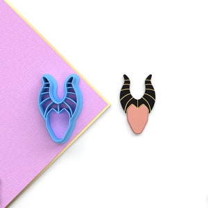 May include: A blue plastic cookie cutter in the shape of Maleficent's head with horns. The cutter is on a pink surface. A black and pink plastic cookie cutter of Maleficent's head with horns is on a white surface.