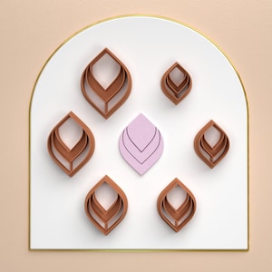 Op de afbeelding: Zeven bruine, gelaagde, bladvormige wanddecoraties zijn in een halve cirkel op een witte achtergrond met een gouden rand gerangschikt. Een enkel, roze, gelaagd, bladvormig stuk bevindt zich in het midden.