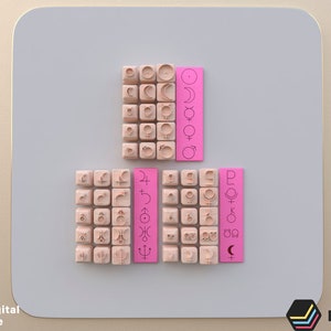 Op de afbeelding: Drie sets houten blokken met hemelse symbolen en roze rechthoekige labels. De blokken zijn gerangschikt in een rasterpatroon. De labels hebben verticale tekst en extra symbolen. De afbeelding heeft een digitaal bestand watermerk.