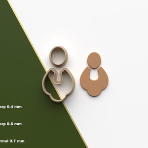 Puede incluir: Dos cortadores de galletas con forma de pendientes, uno de color beige y otro con contorno beige. Los cortadores están sobre una superficie blanca con una franja diagonal verde y dorada. La imagen también incluye texto y un logotipo.