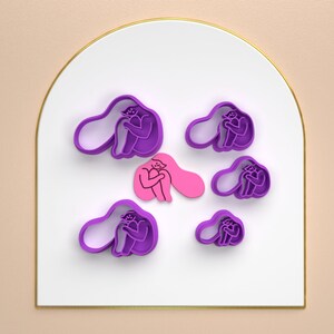 Puede incluir: Un conjunto de siete cortadores de galletas de color morado con forma de personas abrazándose. Los cortadores están sobre una superficie blanca.