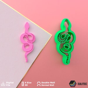 May include: Two 3D printed snake cookie cutters, one pink and one green. The cutters are in the shape of a snake curled up. The cutters are on a pink and white background.