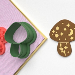 Pode incluir: Um conjunto de dois cortadores de biscoitos, um vermelho e outro verde. O cortador verde tem a forma de um cogumelo com uma parte superior arredondada e um caule. O cortador vermelho tem a forma de uma estrela com um desenho de estrela de cinco pontas.