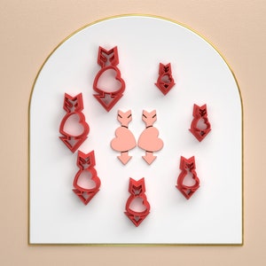 Puede incluir: Un conjunto de ocho cortadores de galletas rojos en forma de flechas y corazones. Los cortadores están dispuestos en círculo sobre una superficie blanca.
