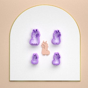 May include: Five purple cookie cutters shaped like llamas, one is a light pink color.