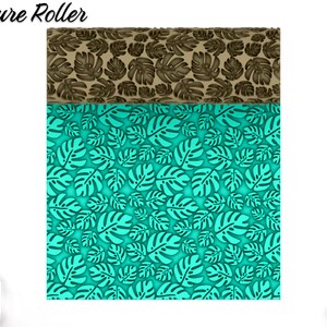 Puede incluir: Un rodillo de textura azul turquesa y marrón con un patrón repetitivo de hojas tropicales. El rodillo está diseñado para crear patrones en relieve en arcilla, fondant u otros materiales de artesanía. "Texture Roller" está escrito en negro en la parte superior de la imagen.