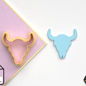 Puede incluir: Un archivo STL impreso en 3D de color dorado de un cortador de galletas con forma de cráneo de toro. El cortador de galletas se muestra junto a un cráneo de toro de plástico azul claro.