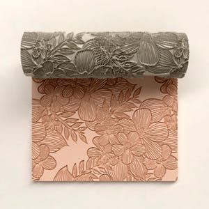 May include: A decorative paint roller with a floral pattern, shown with a matching embossed design on a pink surface. The roller is gray and features a detailed relief of flowers and leaves, creating a textured effect.