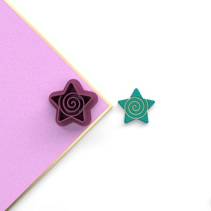 May include: Two star-shaped cookie cutters, one is pink with a gold spiral design and the other is teal with a gold spiral design.