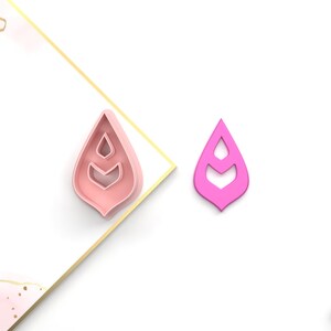 May include: A pink plastic cookie cutter in the shape of a stylized leaf with three layers. The cookie cutter is on a white surface.