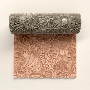 Könnte beinhalten: Eine dekorative Teigrolle mit einem komplizierten floralen und geometrischen Muster. Die Rolle ist grau, und der ausgerollte Abdruck hat eine warme, rötlich-braune Farbe. Das Design weist detaillierte Texturen und Formen auf.