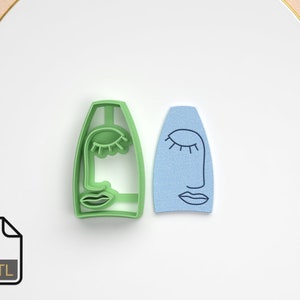 Puede incluir: Dos cortadores de galletas con forma de caras abstractas con los ojos cerrados y una línea para la nariz. Uno es verde y el otro es azul. Están sobre una superficie blanca.