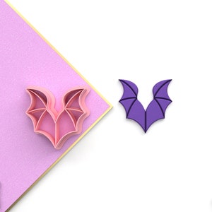 Puede incluir: Un cortador de galletas de ala de murciélago impreso en 3D en rosa y morado. El cortador está diseñado para crear una forma de ala de murciélago para galletas u otros productos horneados.