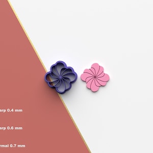 Puede incluir: Dos cortadores de galletas con forma de flor, uno morado y otro rosa, se muestran sobre un fondo blanco y rojo. El cortador morado está a la izquierda y el rosa a la derecha. Los cortadores tienen un diseño en espiral.