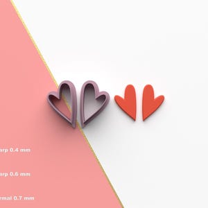 Puede incluir: Dos cortadores de galletas en forma de corazón, un par de color lila claro y otro par de color naranja brillante. El fondo está dividido diagonalmente con secciones rosas y blancas, separadas por una línea dorada.