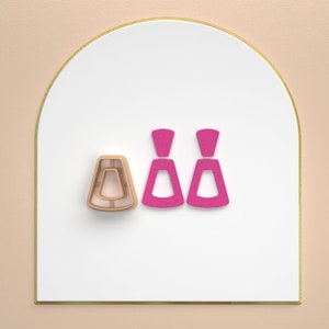 Könnte beinhalten: Zwei geometrisch geformte Ohrringe in Pink und eine goldene Ohrringform auf weißem Hintergrund.