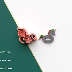 Puede incluir: Dos cortadores de galletas con forma de unicornio. Uno es de color coral, el otro es gris con un cuerno amarillo y una cola arcoíris. Los cortadores están sobre un fondo blanco con una diagonal verde y dorada.