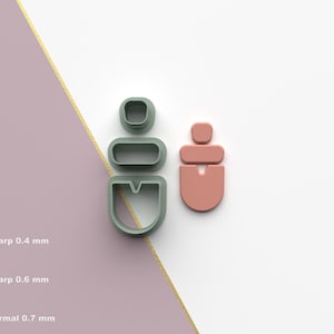 Puede incluir: Dos cortadores de galletas con forma de pendientes, uno en verde salvia y otro en color coral apagado, se muestran sobre un fondo blanco y malva. Los cortadores tienen formas geométricas. La imagen también incluye texto y un icono de archivo STL.
