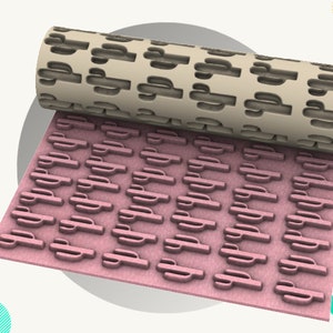 以下が含まれることがあります： ベージュの3Dプリントされたローリングピンで、サボテンのパターンが繰り返されています。ローリングピンは、同じサボテンのパターンがエンボス加工されたピンクの表面を転がっています。画像の左上隅に「STL FILE」というテキストが表示されています。