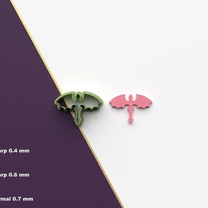 May include: Two dragon cookie cutters, one green and one pink. The cutters are shown on a white surface with a purple background.