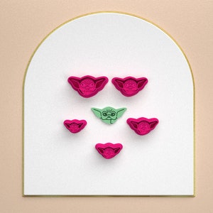 May include: Five cookie cutters in the shape of a character from the Star Wars franchise. The cutters are pink and green. The character is a small, green alien with large ears and a big head.