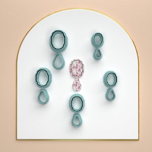 Puede incluir: Conjunto de siete cortadores de galletas azules y blancos en forma de lágrima y óvalos. Los cortadores están dispuestos de forma simétrica sobre una superficie blanca.