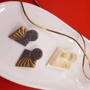 Peut inclure: Une paire de boucles d'oreilles grises et dorées avec un motif géométrique. Les boucles d'oreilles sont sur une assiette blanche avec un ruban doré. Il y a un moule en plastique blanc pour faire les boucles d'oreilles.