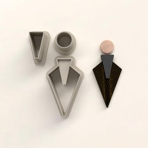 Puede incluir: Un conjunto de componentes de pendientes y un pendiente terminado. Los componentes son de plástico gris claro, incluyendo un cortador en forma de V, un círculo y un cortador en forma de diamante. El pendiente terminado tiene un círculo rosa, un triángulo gris y un diamante con estampado negro y dorado.