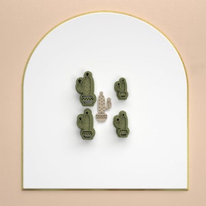 Puede incluir: Cinco cortadores de galletas de cactus verde sobre una superficie blanca con un borde dorado. Los cortadores de galletas tienen la forma de una planta de cactus con una maceta.