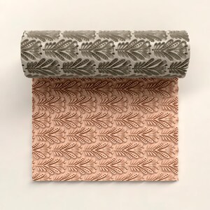 Puede incluir: Un rodillo decorativo con un patrón de hojas repetido. El rodillo es gris y el patrón en relieve es visible en una hoja de material rosa. El patrón presenta hojas estilizadas con detalles punteados.