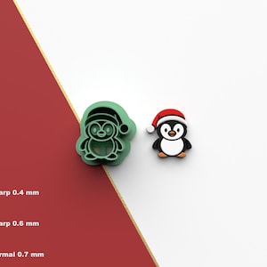 Pode incluir: Um cortador de biscoitos verde em forma de pinguim e uma pequena figura de pinguim usando um gorro de Papai Noel. O cortador de biscoitos é verde, e a figura é preta, branca e vermelha. O fundo é branco e vermelho.