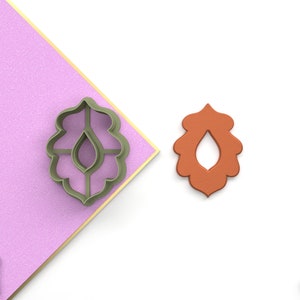 Puede incluir: Un cortador de galletas de plástico verde en forma de hoja estilizada con un agujero en forma de lágrima en el centro. Un cortador de galletas de plástico marrón de la misma forma se muestra al lado.