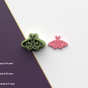 May include: Two moth-shaped cookie cutters, one green and one pink, are displayed on a white surface. The green cutter has intricate details, while the pink one is solid. The image also shows text indicating different cutting edge thicknesses.