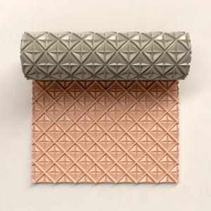 May include: A rolled grey patterned object sits on top of a flat, unfolded, peach-coloured patterned surface. Both surfaces have a repeating geometric design of diamond shapes with star-like details, creating a textured appearance.