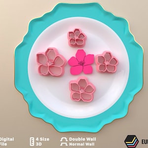 Puede incluir: Cinco cortadores de galletas de plástico rosa y blanco con forma de flores de cerezo en un plato blanco con borde turquesa. Los cortadores son de diferentes tamaños.