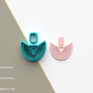 Puede incluir: Dos cortadores de galletas con forma de pendientes. Uno es turquesa, el otro rosa. Cada uno tiene una parte superior rectangular y una parte inferior en forma de abanico con un triángulo en el centro. El cortador turquesa tiene líneas finas que irradian desde el triángulo.