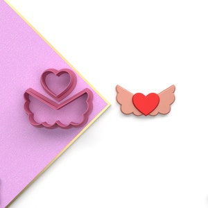 Puede incluir: Dos cortadores de galletas de plástico rosa en forma de alas de ángel con un corazón en el centro. Las alas están sobre un fondo rosa y el corazón está sobre un fondo blanco.