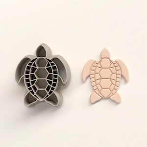 Pode incluir: Dois cortadores de biscoitos em forma de tartaruga. Um é cinza e tem um padrão de grade dentro da concha. O outro é uma forma de tartaruga rosa claro sólida. Ambos têm barbatanas e cabeças detalhadas.
