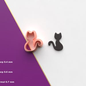 Puede incluir: Un cortador de galletas rosa con forma de gato y un objeto negro con forma de gato se muestran sobre un fondo blanco y morado. El cortador tiene una cola rizada. La imagen también muestra el icono del archivo STL.