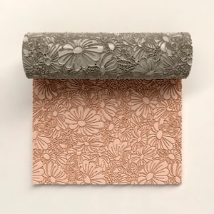 Könnte beinhalten: Eine graue Blumenmusterwalze mit einem passenden geprägten Design auf einer rosa Oberfläche. Die Walze ist so konzipiert, dass sie ein strukturiertes Blumenmuster erzeugt. Das Design zeigt Gänseblümchen und andere florale Elemente.
