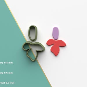 Puede incluir: Dos cortadores de galletas impresos en 3D, uno verde y otro rojo. El cortador verde tiene forma de hoja con un tallo redondeado. El cortador rojo tiene forma de flor con cinco pétalos.