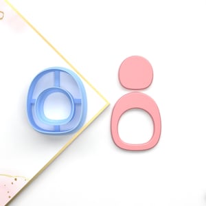 Puede incluir: Dos cortadores de galletas, uno azul claro y otro rosa, con forma de la letra "O". El cortador azul claro tiene un círculo más pequeño dentro del círculo más grande. El cortador rosa tiene un círculo más pequeño dentro del círculo más grande.